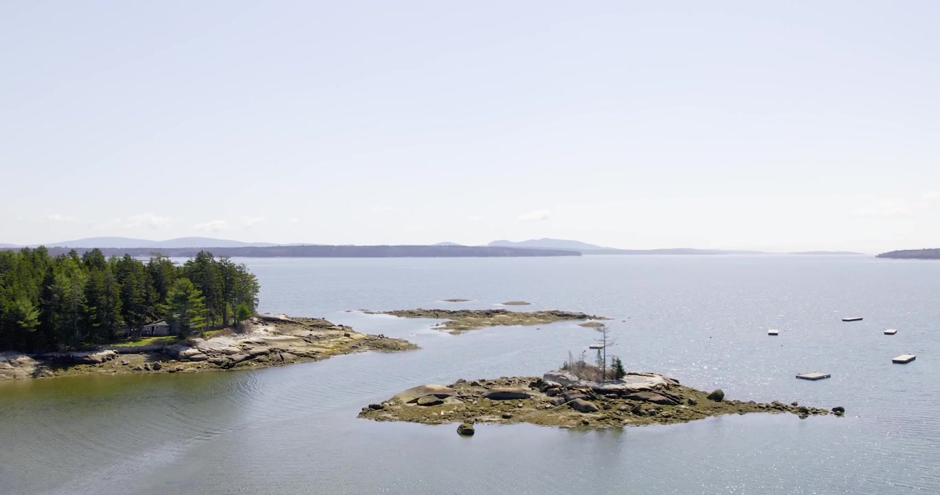Sebuah pemandangan udara luas menunjukkan pantai Maine yang tenang dengan pulau-pulau, pepohonan, dan perairan terbuka di siang hari yang cerah.