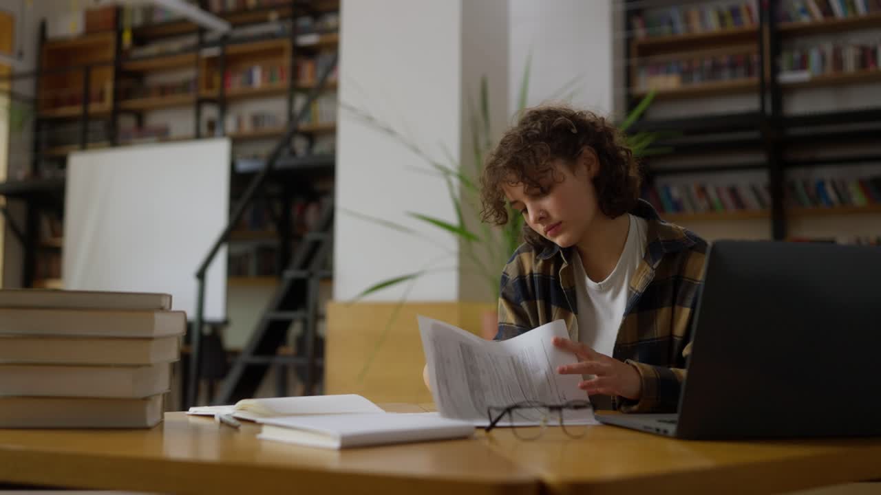 concentrada chica morena en una camisa a cuadros con cabello rizado se sienta en la mesa y trabaja en la biblioteca