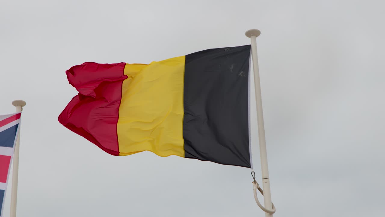Belgian and British flags wave vigorously on flagpoles, overcast coastal sky, steady camera, natural light