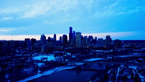 Winter-evening-downtown-Edmonton-festive-LED-lights-on-high-rises-as-drone-reverse-fly's-backward-and-downwards-snow-covered-parks-buildings-by-the-98-Ave-NW-James-Macdonald-and-the-Low-Level-Bridge