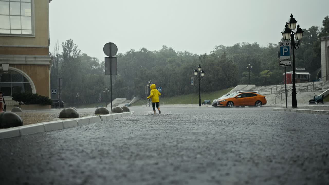 황색 재을 입은 행복한 십대 소녀의 뒷면은 많은 비가 내리는 동안 공원에서 웅이와 물을 가로질러 달리고 있습니다.