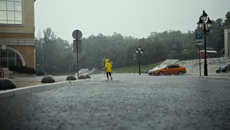 vista trasera de una adolescente feliz en una chaqueta amarilla corriendo a través de charcos y agua en el parque durante una fuerte lluvia