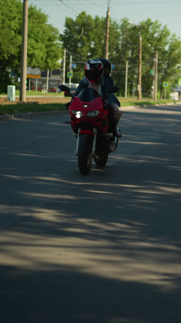 dos hermanas montan una bicicleta eléctrica roja, ambas con cascos, en una carretera sombreada, se ve un coche arrancando por detrás mientras otro se mueve en el carril opuesto, los árboles alinean la carretera