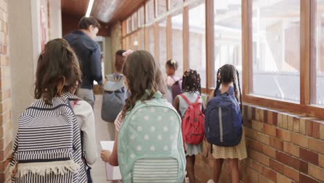 Feliz-Y-Diverso-Maestro-Y-Niños-Con-Bolsas-Caminando-En-El-Corredor-De-La-Escuela-Primaria,-Cámara-Lenta