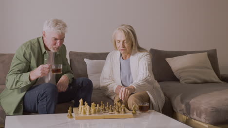 Senior-Man-And-Woman-Playing-Chess-And-Drinking-Tea-While-Sitting-On-Sofa-At-Home