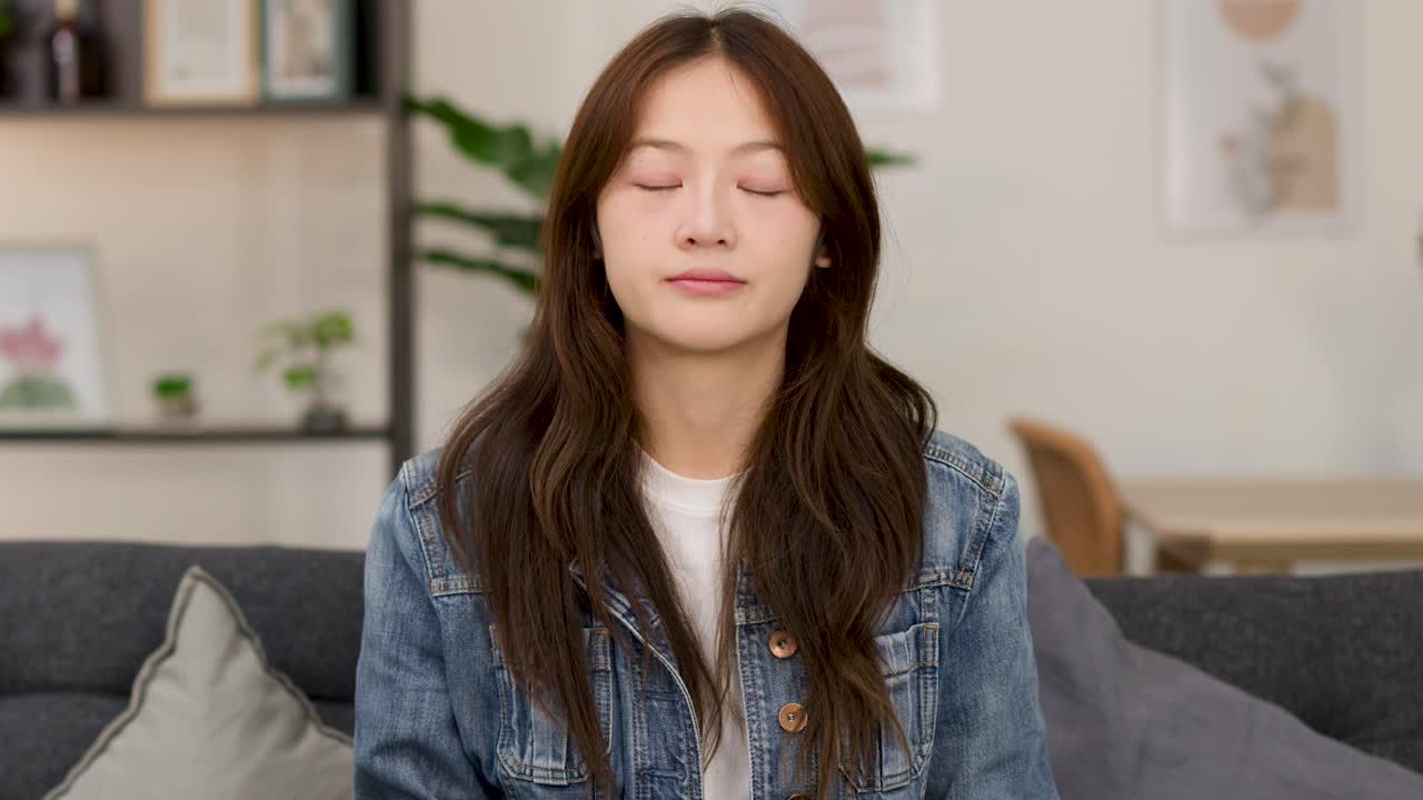 Casual young woman shrugs with uncertain expression on sofa, static camera, soft indoor lighting