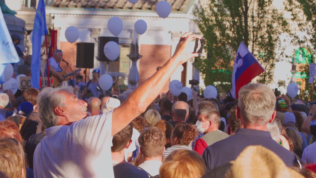 Old Man Taking Video Of Audience Watching Band Performers On Stage During Statehood Day In Ljubljana, Slovenia. medium shot