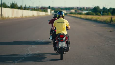 Instructor-De-Manejo-En-Un-Ciclomotor-Con-Una-Camiseta-Amarilla-Sigue-A-Su-Estudiante-Con-Una-Camiseta-Roja-En-Una-Carretera-Ancha