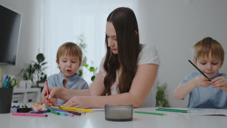 Sitting-at-the-table-in-the-living-room,-a-young-mother-teaches-her-son-to-hold-a-pencil-and-teaches-to-draw