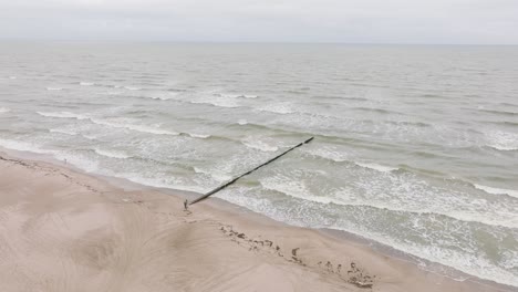 establishing aerial view of baltic sea coast on a overcast day, old wooden pier, white sand beach, large storm waves crushing against coast, climate changes, wide drone shot moving forward tilt down