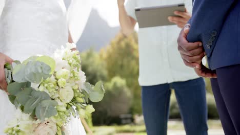 Sección-Media-De-Un-Hombre-Diverso-Con-Una-Tableta-Oficiando-El-Matrimonio-De-Una-Pareja-En-Un-Jardín-Soleado-En-Cámara-Lenta