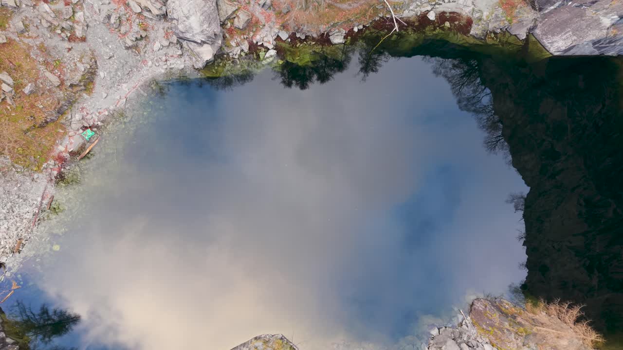 A drone ascends above a quarry lake that mirrors blue sky and drifting clouds, gradually revealing the rocky edges and towering slate walls in a hypnotic upward view