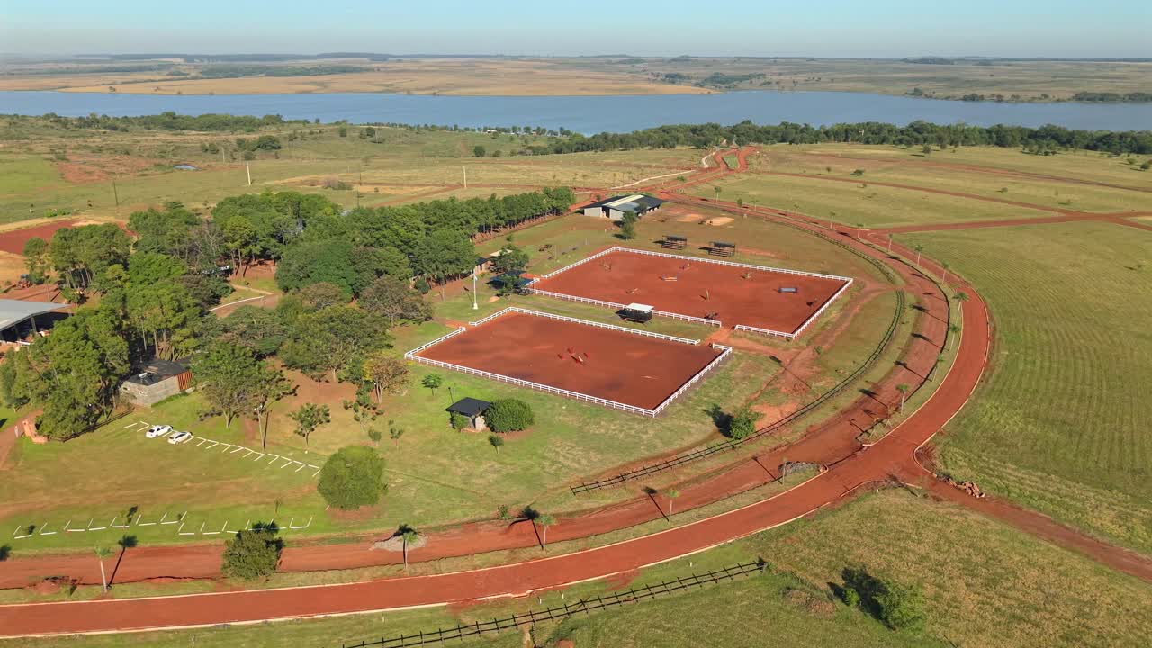 Smooth sideways drone shot captures equestrian club’s pristine arena, flanked by golden fields and winding river under vast skies. Ideal for luxury sports or property showcases