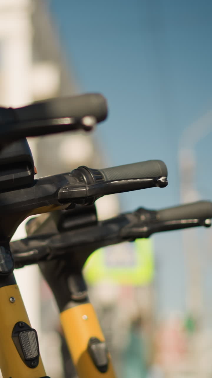 Flota de scooters a la luz del sol alineada en la acera, composición nítida a la luz del día con tallos amarillos y cielo azul despejado, múltiples manillares y empuñaduras enfocados, telón de fondo de calle urbana, viajeros escaneando la app, alquiler