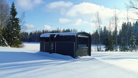 Aerial-drone-backward-moving-shot-over-a-rectangular-cabin-surrounded-by-white-snow-covered-landscape-and-a-frozen-lake-in-the-background