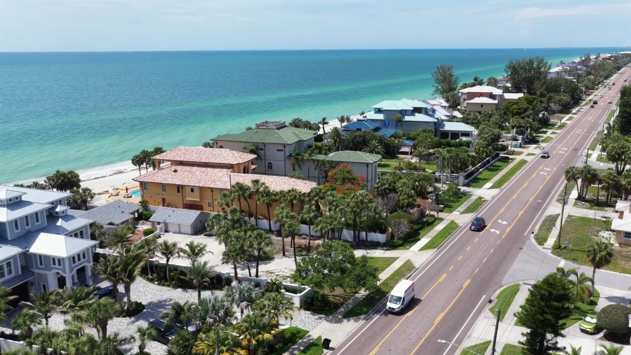 Lyxiga strandvillor kantar en lugn kustväg i Indian Rocks Beach, Florida, där turkosa golfvatten möter vit sandstrand intill palmbeskuggade uppfarter och pooler. Flygfoto med drönare.