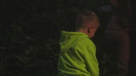 child crashes through thick bushes in night park. little boy follows sister walking across dark woods at nighttime. boy and girl adventures in forest