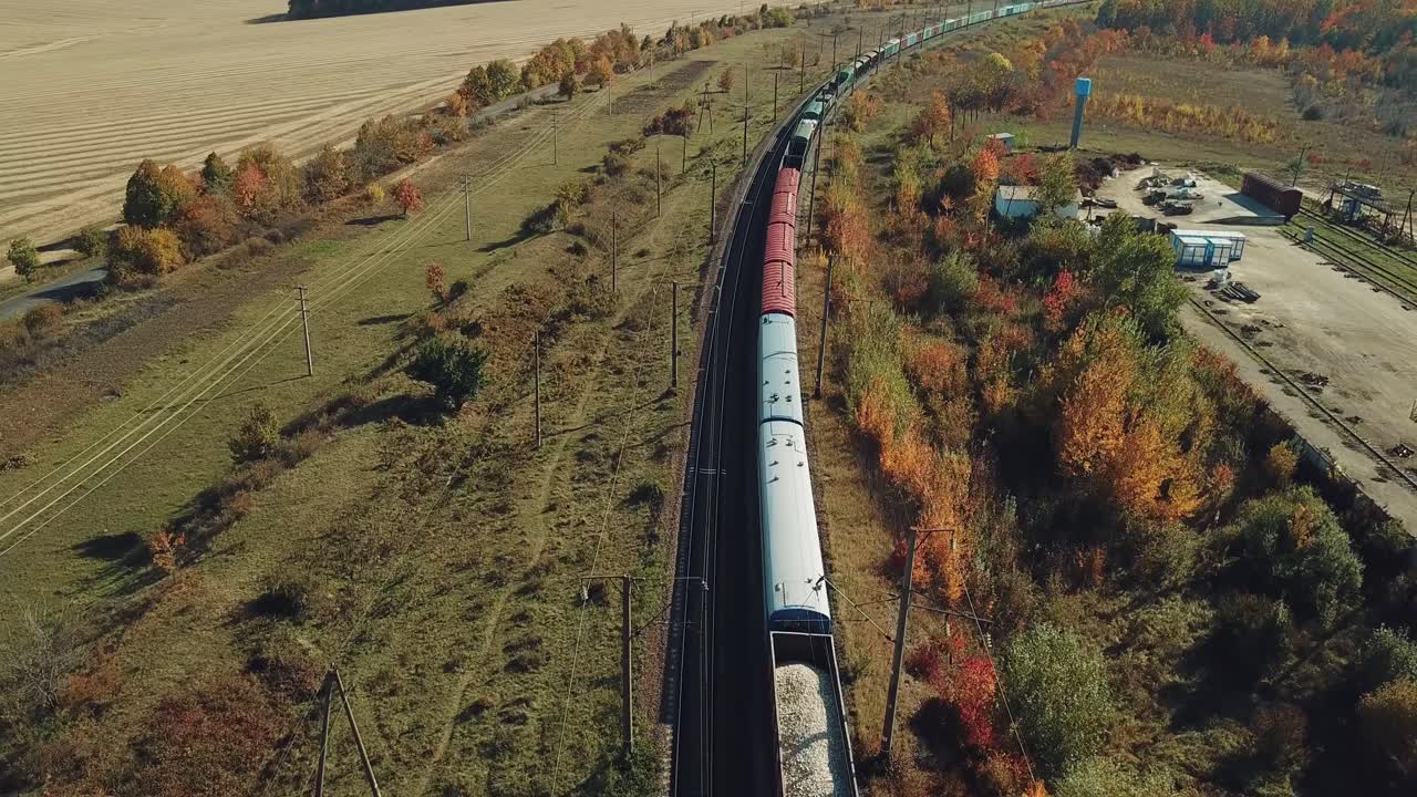 a long freight train with building materials is moving by railway on the background of fields and forests in the summer on a sunny day. Aerial view