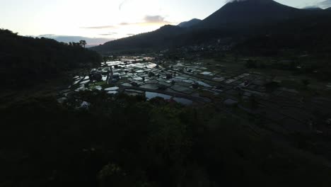 Cinematic-drone-shot-descending-into-a-valley-with-expansive-rice-fields,-with-a-volcano-in-the-background,-all-bathed-in-the-golden-hour-light