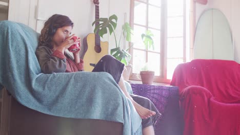 Relaxed-mixed-race-woman-wearing-headphones-sitting-drinking-tea-in-sunny-cottage-living-room