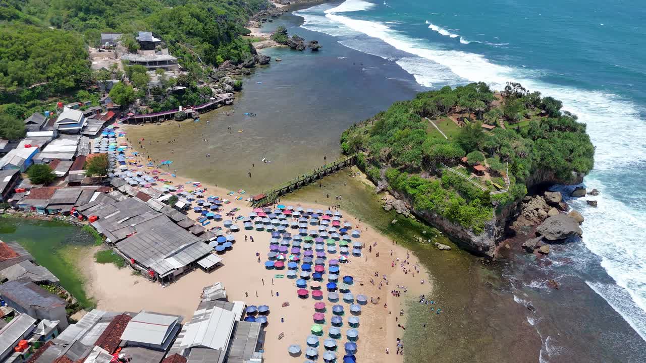 Aerial drone view of Drini Beach in Yogyakarta, Indonesia, showing colorful umbrellas, visitors enjoying the shallow water, and a small green island surrounded by ocean waves