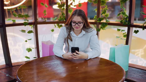 mujer joven sonriente sentada en una mesa de madera usando un teléfono inteligente, rodeada de bolsas de compras, plantas verdes enmarcan la escena, mientras que las luces de neón con temas de gatos en el fondo