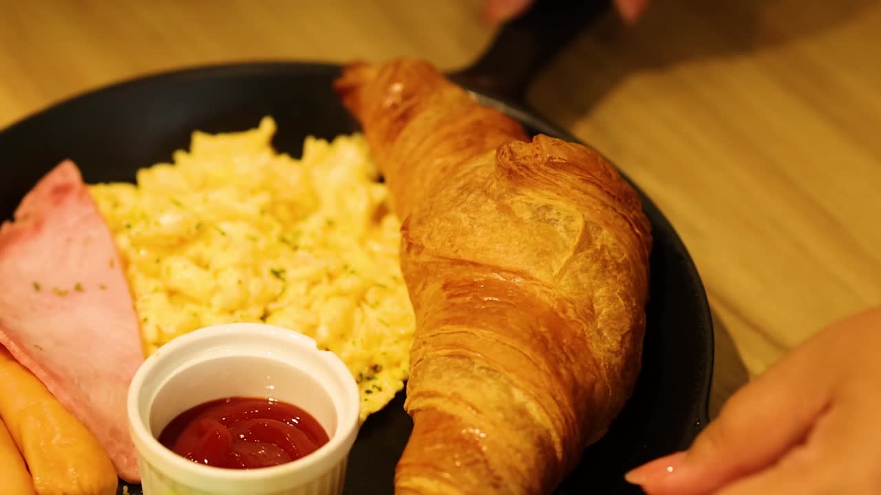 A close-up of a breakfast plate featuring scrambled eggs, croissant, ham, and ketchup.