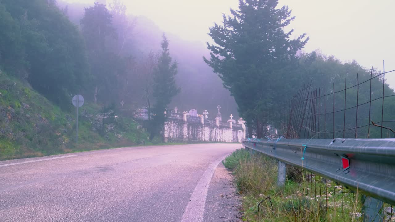 Static shot of wet road turn ending in a cemetery, Nature and woods around it. A bit of wind. Foggy weather.