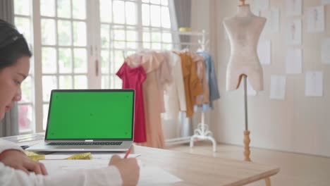 diseñadora asiática dibujando en papel con una pantalla verde en una computadora portátil