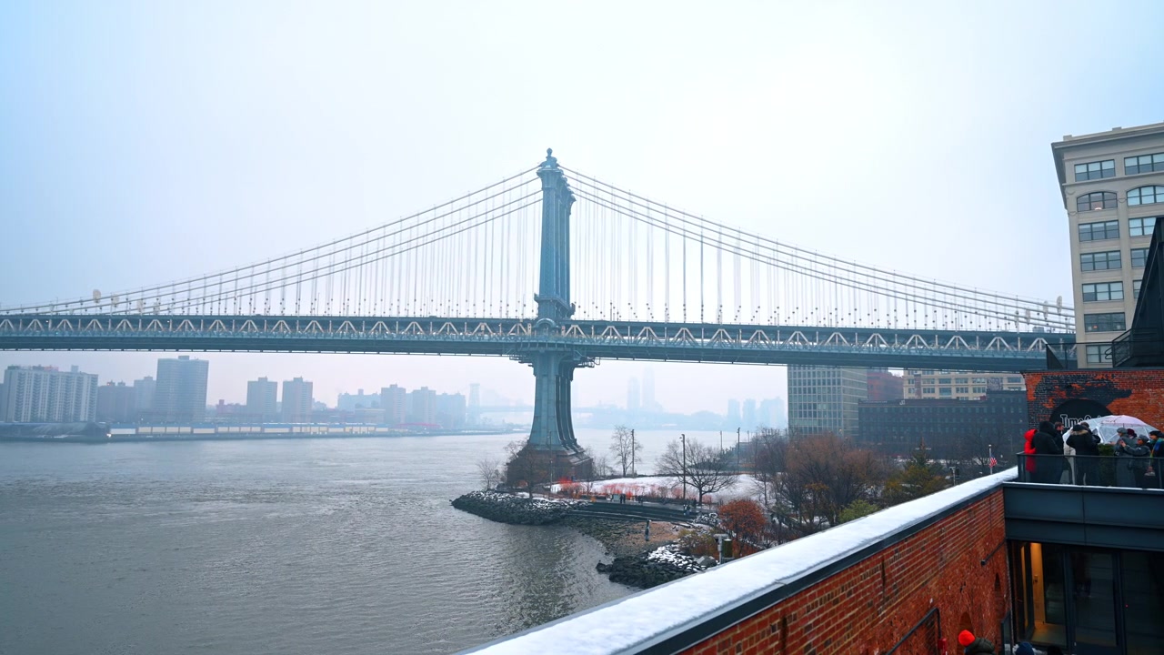 New York, USA, 25 dicembre 2025: Vista invernale nuvolosa del Ponte di Manhattan, New York City, New York. Persone si radunano su una terrazza rialzata in mattoni che si affaccia sul fiume in una giornata nuvolosa