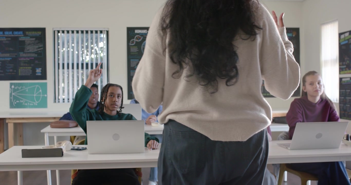 Indian teacher teaching teenagers front of room after eye contact, student raising hand, laptops