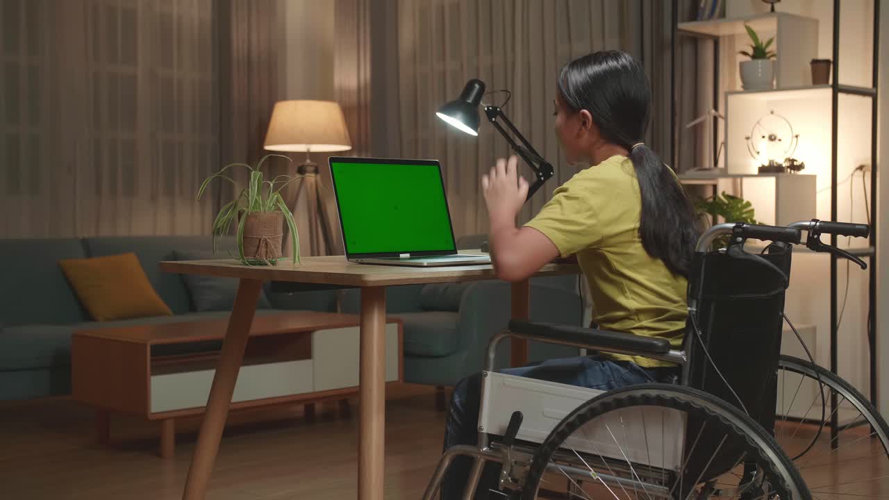 Asian Kid Girl Sitting In A Wheelchair And Raise Hand Before Asking A Question While Learning Online On Laptop Computer With Mock Up Green Screen At Home