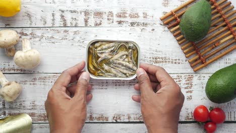 mano sosteniendo una lata de anchoas en una mesa de madera con otros ingredientes