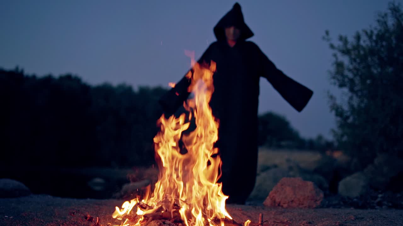 Fire and black figure at darkness. Spooky death in cloak with hood standing against the bright flame and doing some ritual outdoors. Halloween.
