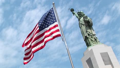 Die-Freiheitsstatue-Steht-Majestätisch-Auf-Ihrem-Sockel,-Während-Im-Vordergrund-Eine-Amerikanische-Flagge-Weht