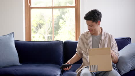 Happy-biracial-man-opening-delivery-in-cardboard-box-and-using-smartphone,-copy-space,-slow-motion