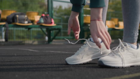 close-up of lady in green sweater loosening shoelace with blurred view of people playing basketball outdoors, emphasizing focus on footwear with active outdoor scene in background