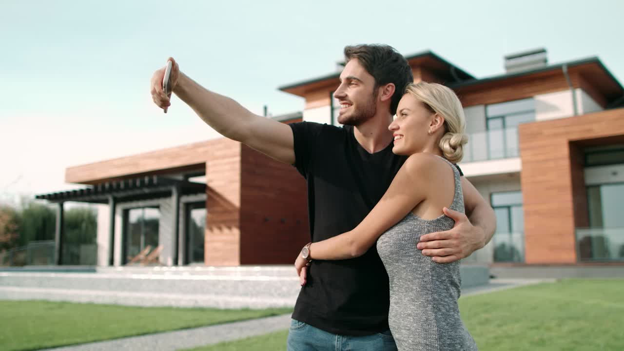 pareja alegre tomando una foto selfie en el teléfono móvil cerca de una casa de lujo.