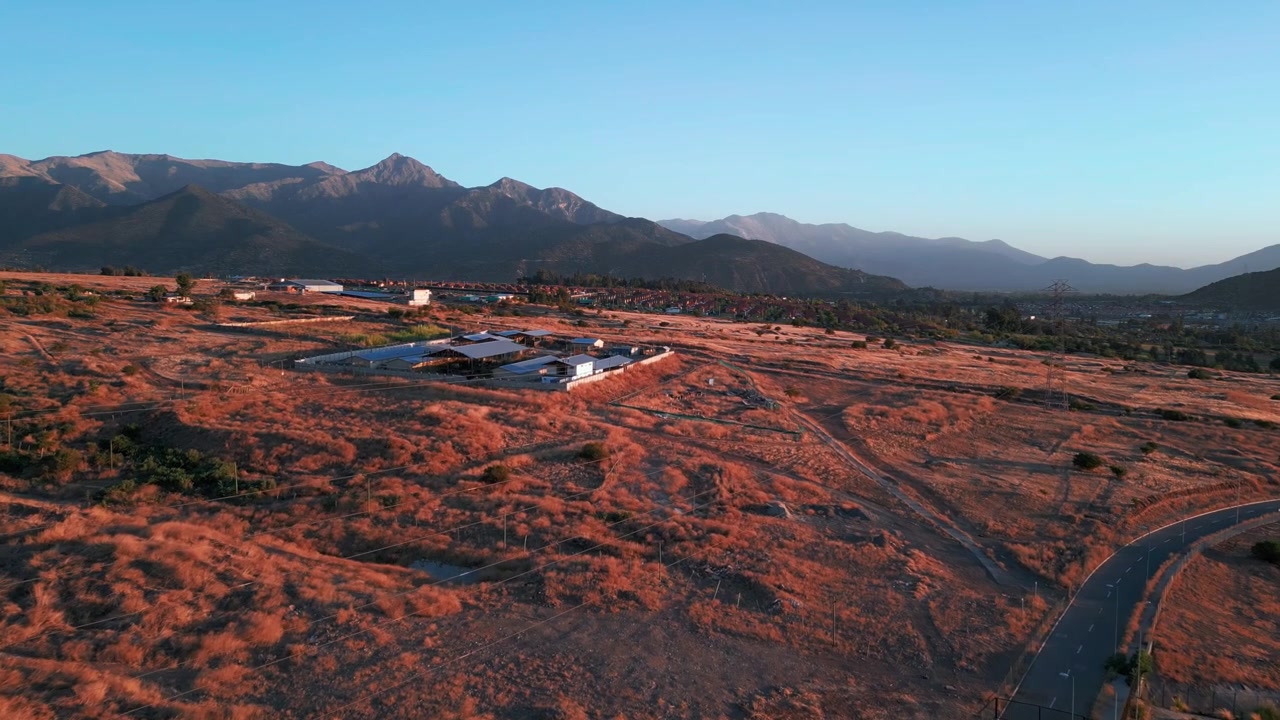 Volo in avanti sopra un terreno arido collinare con terra rossa secca, edifici sparsi e strada tortuosa che rivela una drammatica catena montuosa all'ora d'oro sotto un cielo azzurro limpido