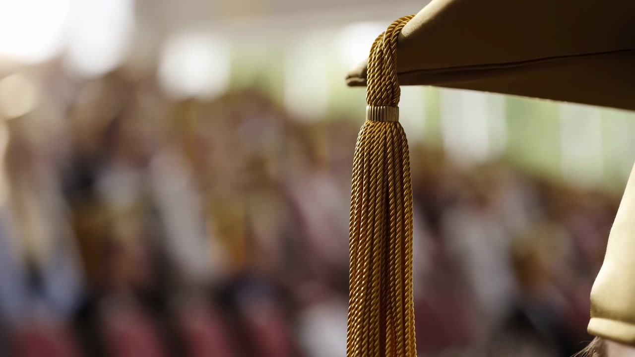 Nærbillede bagfra fra siden af en kandidat iført studenterhue med guldflettet kvast hængende fra hueranden, varmt indendørs lys, sløret publikum, formel ceremoniatmosfære og akademisk feststemning.