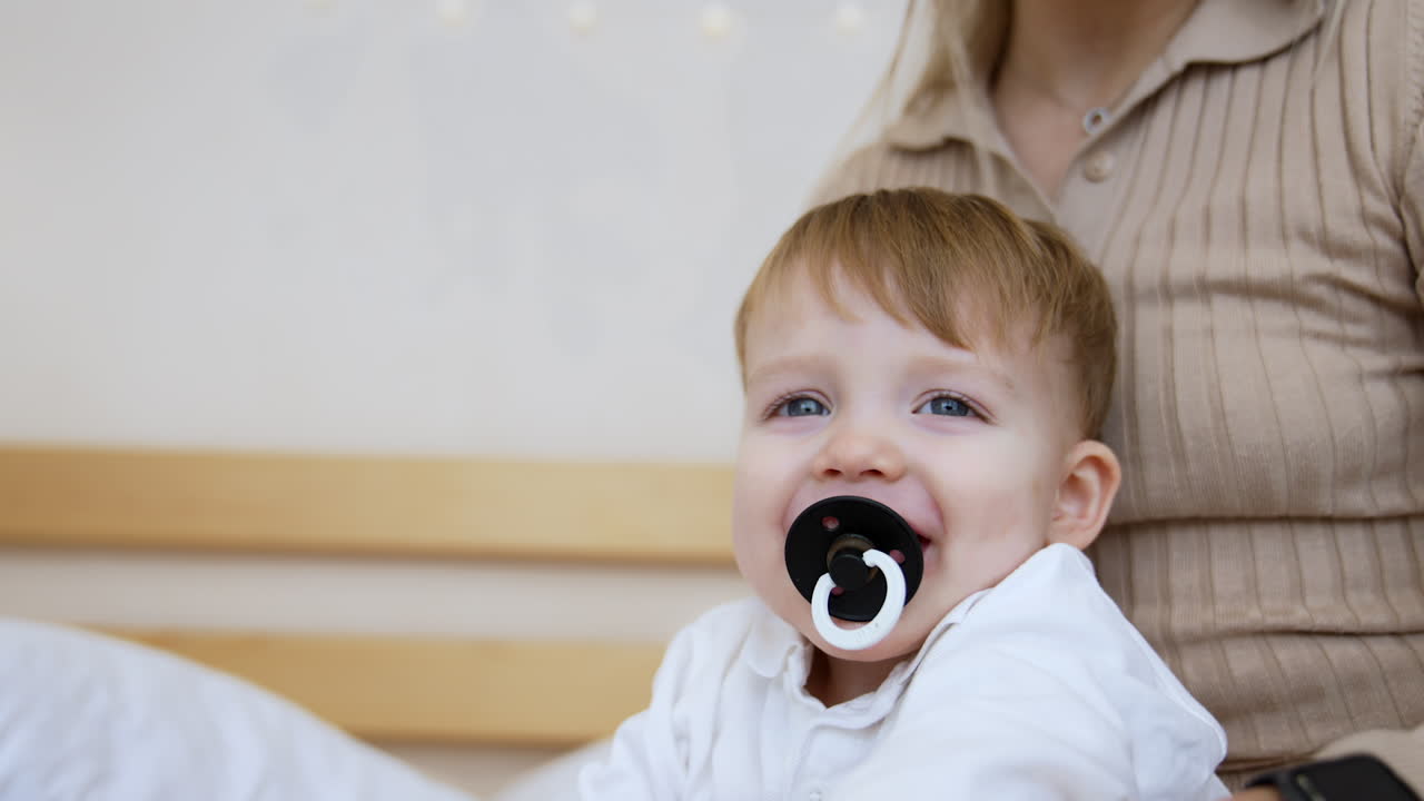 Adorable Caucasian grey-eyed baby boy with a pacifier in mouth. Close up. Kid sits in mom's hands and mother strokes son's palm with a finger.