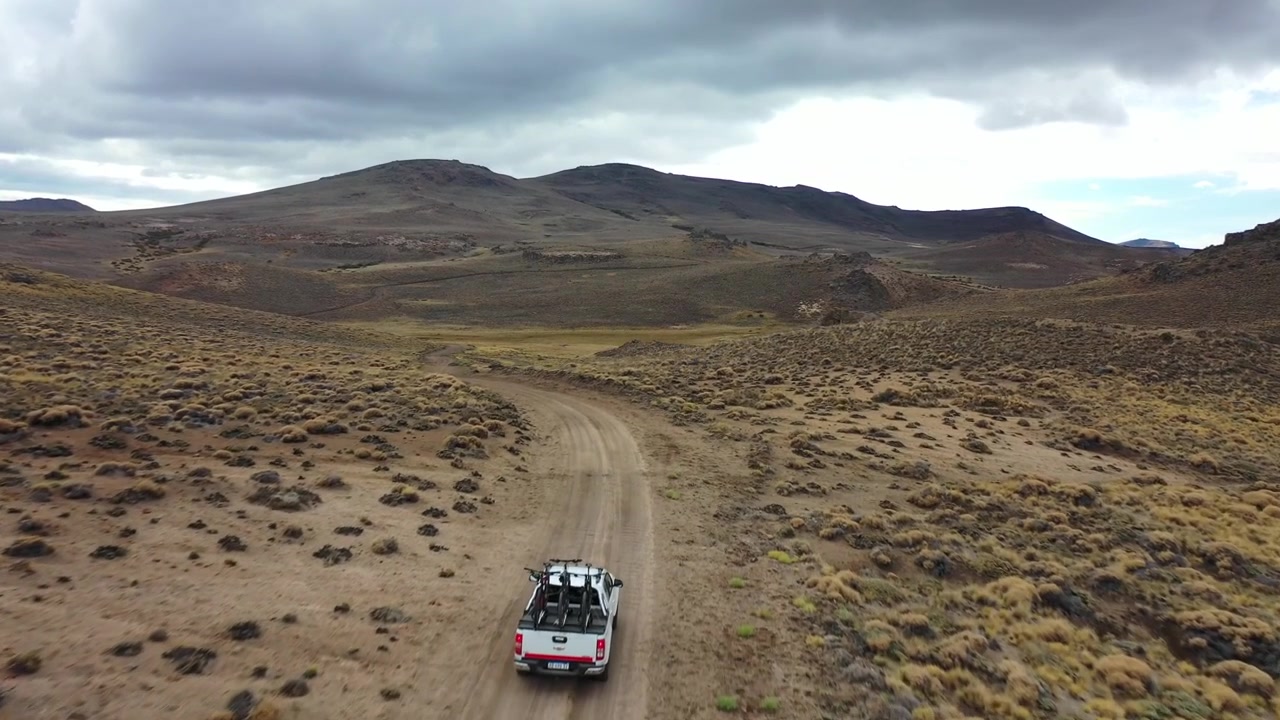 Ripresa con drone dall'alto di un veicolo 4x4 bianco che percorre un sentiero sterrato tortuoso attraverso una vasta valle di montagna a Bariloche, Argentina. Evidenzia viaggi remoti, esplorazione avventurosa e natura selvaggia.