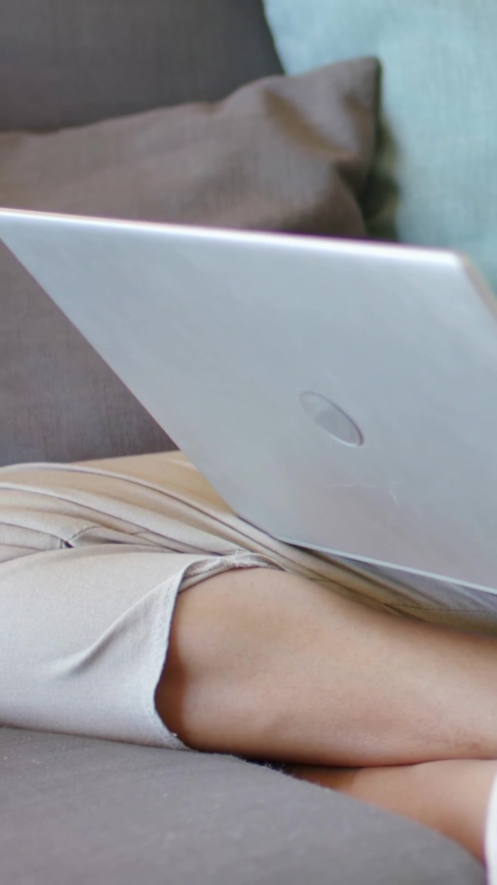 Vertical video: Adjusting woman responding to silver laptop shift on couch at home, wearing skirt