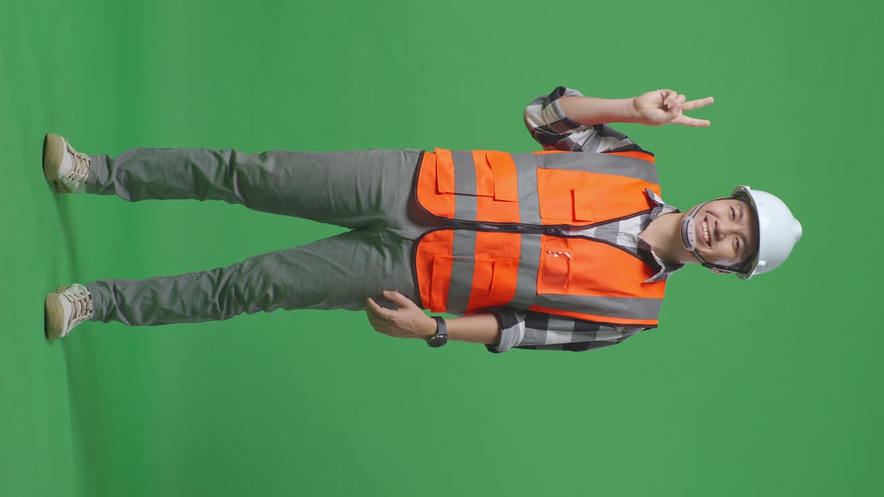 Full Body Of Asian Male Engineer With Safety Helmet Smiling And Showing Peace Gesture While Standing In The Green Screen Background Studio