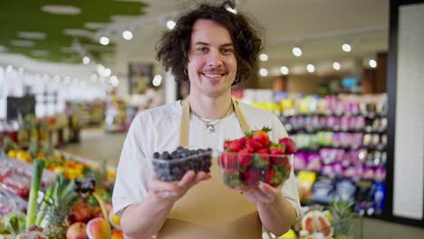 retrato de un tipo feliz con el cabello rizado un trabajador de un supermercado que sostiene en sus manos pequeñas cajas de arándanos y fresas en el supermercado