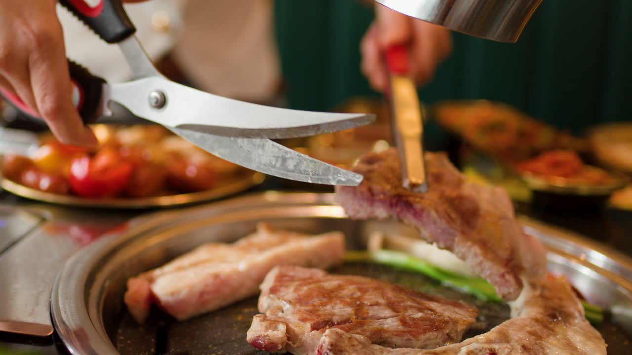 Hands cut sizzling meat on tabletop grill using scissors and tongs, warm lighting, shallow focus
