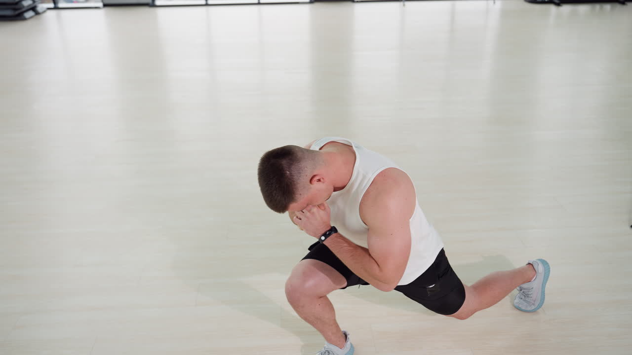Male modern trainer stretching legs in fitness center on wooden floor performing side lunge with clasped hands wearing white tank top black shorts under large windows and overhead lighting