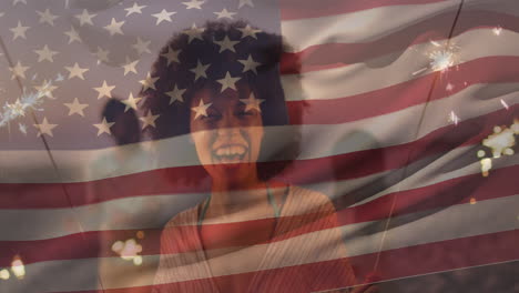 video compuesto de la bandera de los ee.uu. sobre una feliz mujer afroamericana celebrando con sparkler en la playa