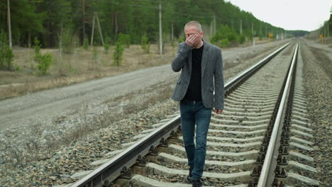 a close-up view of a middle-aged man in a grey suit and black shirt walking along railway tracks in a forested area, the man covers his face with one hand, appearing deep in thought