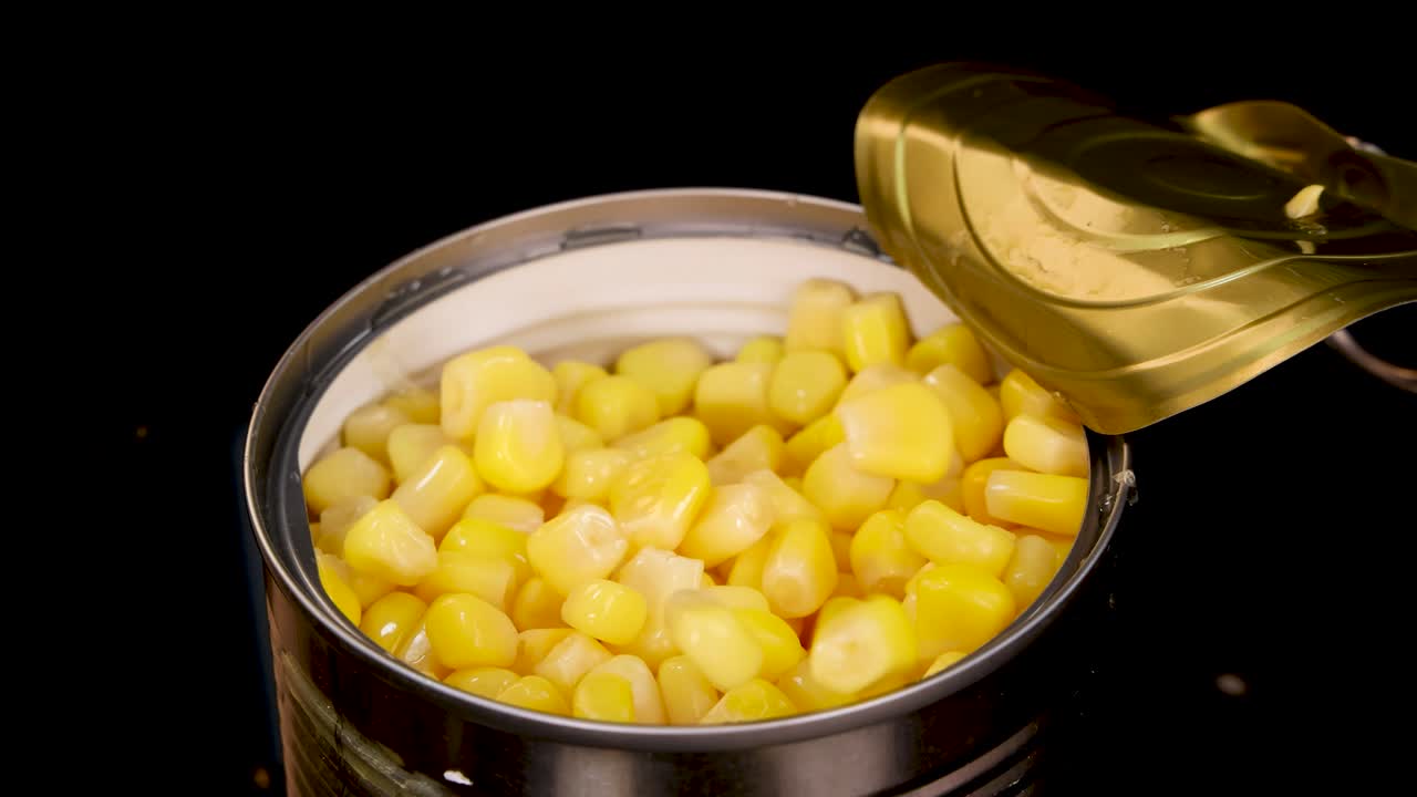 A single kernel of cooked corn falls into an open can of canned corn against a black background, captured in high-speed, well-lit close-up